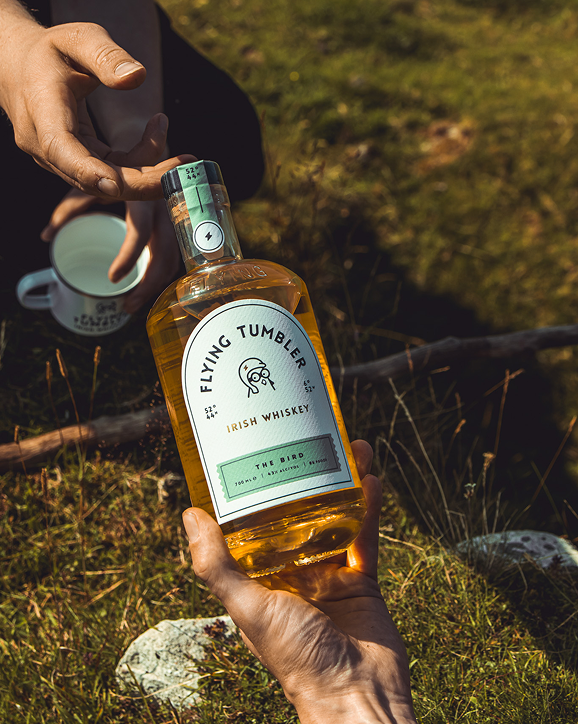 A bottle of Flying Tumbler The Bird Irish whiskey held outdoors while pouring a measure into a mug.