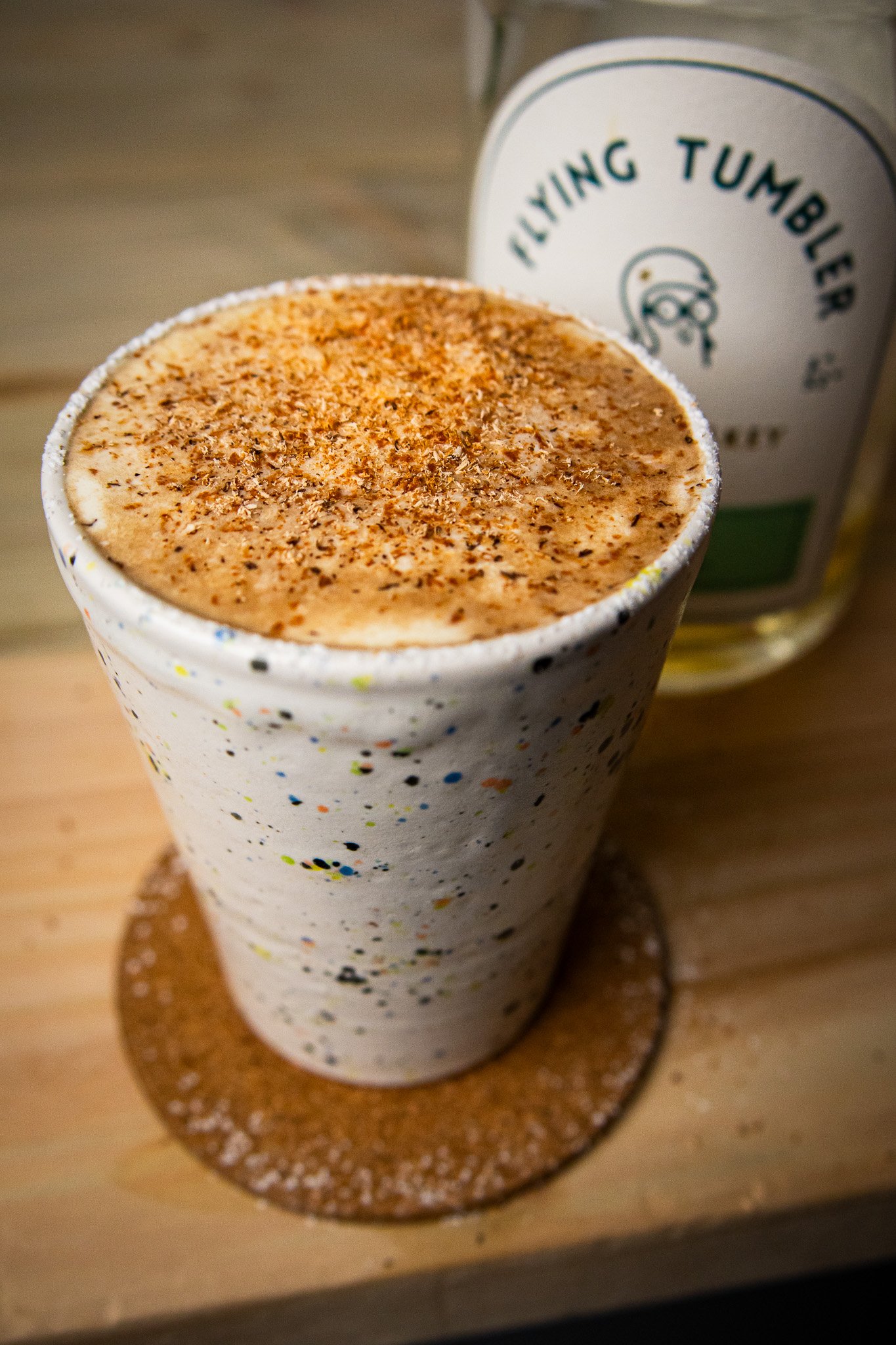 Irish coffee dusted with grated nutmeg in a speckled ceramic tumbler beside a bottle of Flying Tumbler Irish whiskey